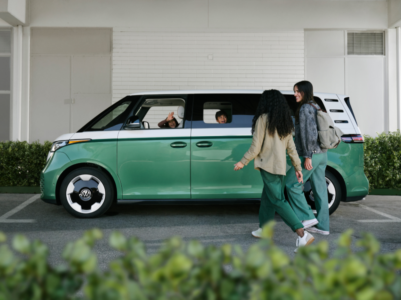 Un ID. Buzz EV mostrado en Mahi Green Metallic y Candy White estacionado frente a un edificio y setos verdes, con dos niños dentro del vehículo saludando alegremente a dos personas que se acercan caminando.
